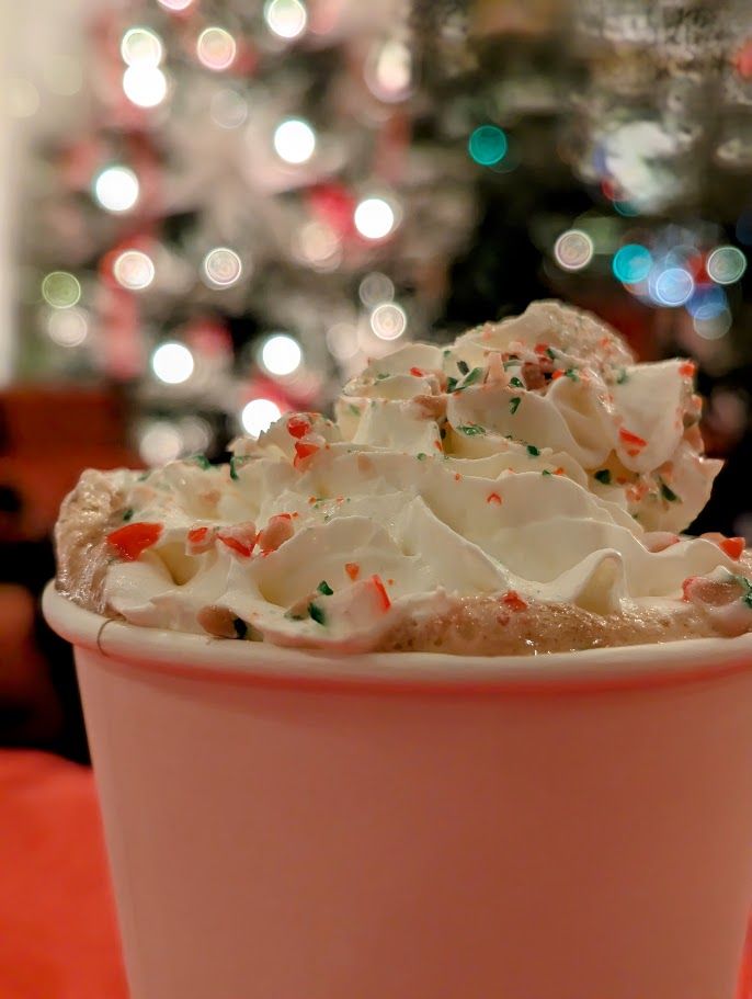 A hot drink topped with whipped cream and crushed candy cane in front of Christmas lights